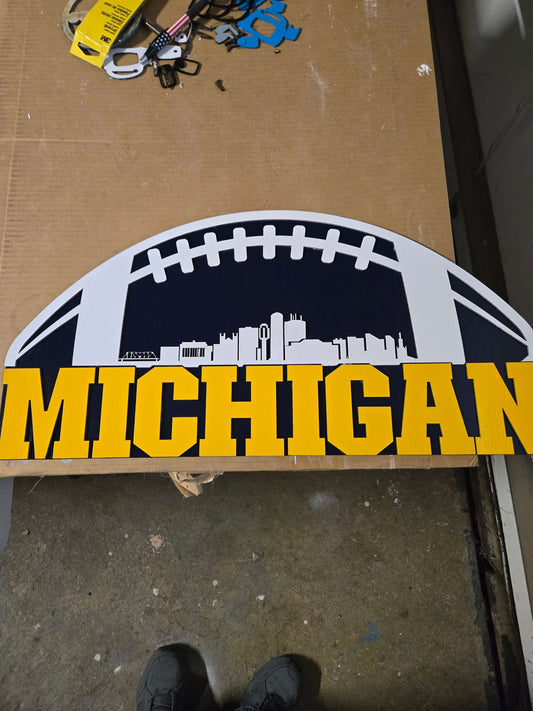 Wooden decor sign featuring the word Michigan with a city skyline background and football shape design.